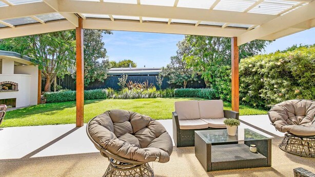 Une terrasse extérieure couverte, aménagée avec des meubles en osier, une table en verre et offrant une vue sur un jardin avec un barbecue et de la verdure.