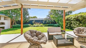 Eine überdachte Terrasse im Freien mit Korbmöbeln, einem Glastisch und Blick auf einen Hinterhof mit Grill und viel Grün.