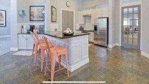 Una cocina amplia con armarios blancos, una isla central con taburetes de bar, un refrigerador de acero inoxidable y un ventanal grande.