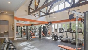 Un gimnasio con diverso equipamiento de ejercicio, amplias ventanas y techo alto.
