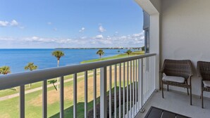 A balcony with wicker chairs, a view of the ocean, and a grassy area with palm trees.