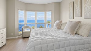 A bedroom with a large bed, a chair, and a view of the ocean through a bay window.