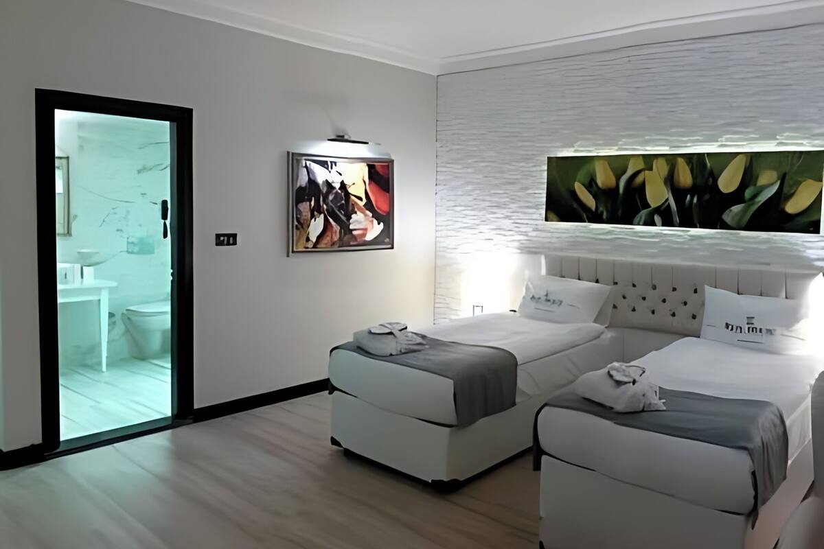 Ein modernes Hotelzimmer mit zwei Betten, einem großen Fenster mit Blick, einem durch eine Glastür einsehbaren Badezimmer und einem an der Wand befestigten Kunstwerk.