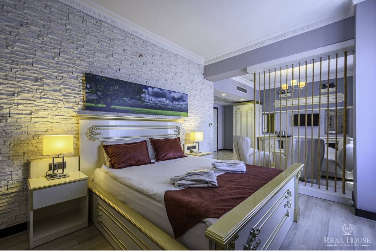 Ein Hotelzimmer mit weißer Ziegelwand, einem großen Landschaftsbild und einem Bett mit roter Tagesdecke, sowie einem Nachttisch mit Lampe.