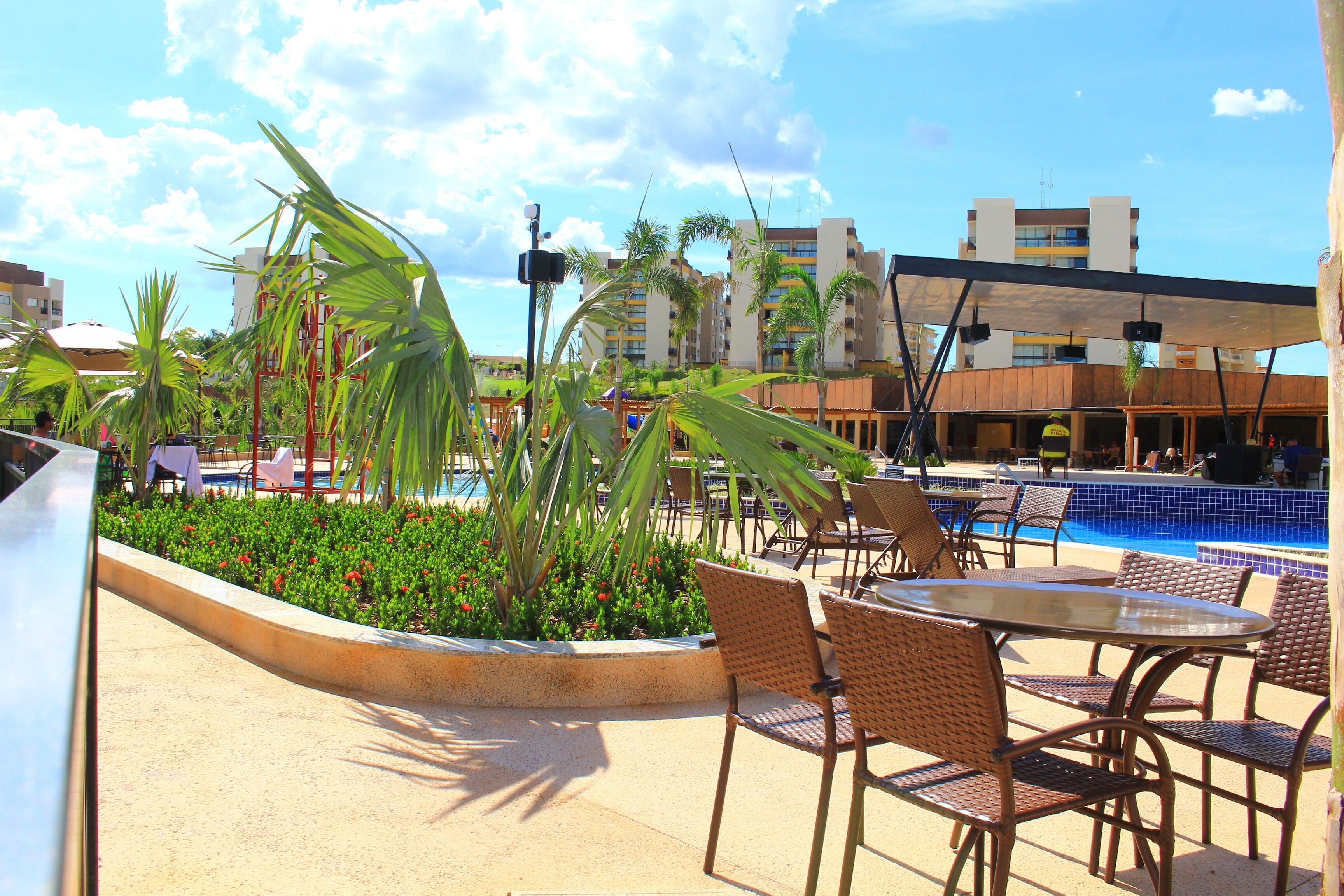 An outdoor pool area with lounge chairs and a dining table, surrounded by palm trees and a clear sky.