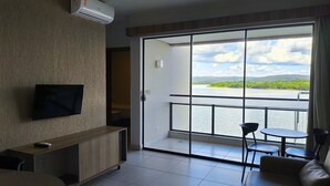 A modern room with a TV on the wall, a wooden console table, a sofa, and a balcony with a view of a lake and greenery. - Praias do Lago Eco Resort by WAM Experience (Caldas Novas)