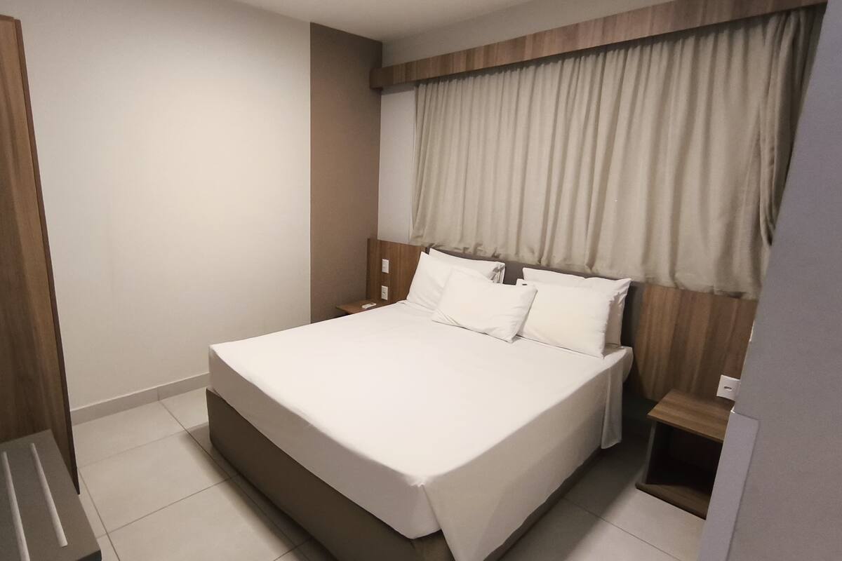 A modern hotel room with a large bed, wooden headboard, and a window with curtains.