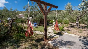 Eine Holzpergola mit roten und weißen Streifen, von der aus schaukelnde Hängematten hängen, eingebettet in einen Garten mit einem Steinweg und Olivenbäumen.