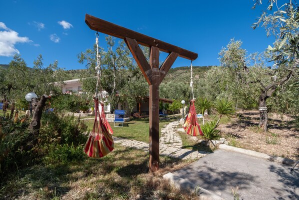 Una pérgola de madera con hamacas a rayas rojas y blancas, ubicada en un jardín con un camino de piedra y olivos.