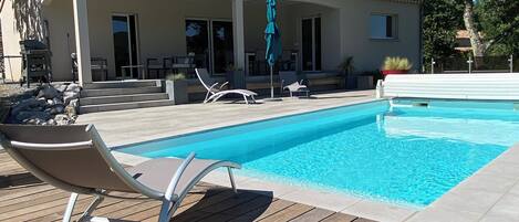 A pool area with a wooden deck, lounge chairs, and a covered patio with outdoor furniture.