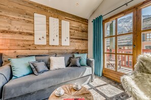 A cozy room with a grey sofa, wooden wall paneling, a large window with a view of mountains, and a fluffy white chair.