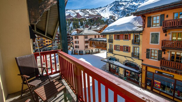 A balcony with wooden chairs and a bench, overlooking a snow-covered mountain and colorful buildings.