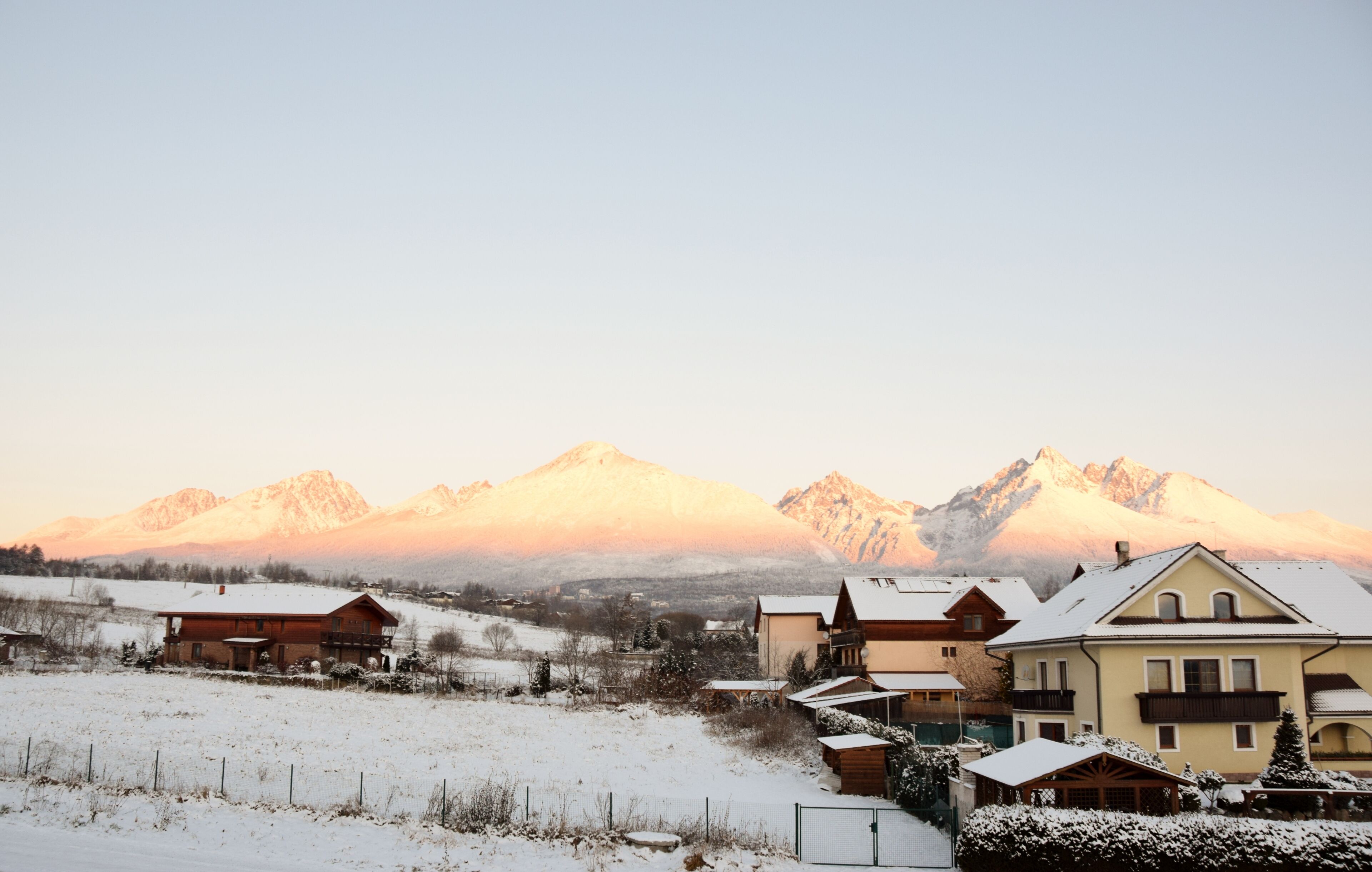 A snow-covered landscape with a mountain range in the background and several buildings with snow-covered roofs.