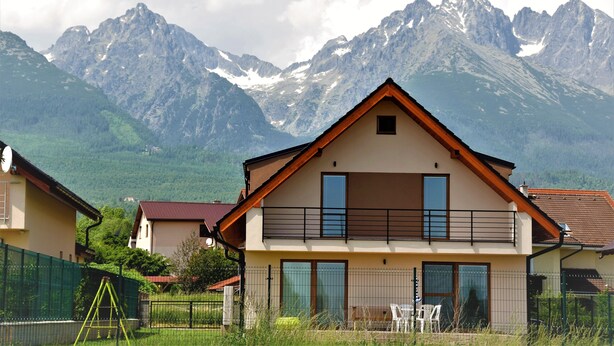 Una casa de dos pisos con un balcón, rodeada de montañas y una valla verde.