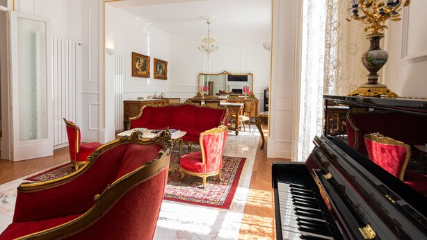 Una sala elegante con un piano, sillas tapizadas en rojo, un candelabro y un espejo.