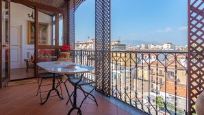 Un balcón con una mesa, sillas, una planta en maceta y vistas a edificios y montañas a lo lejos.
