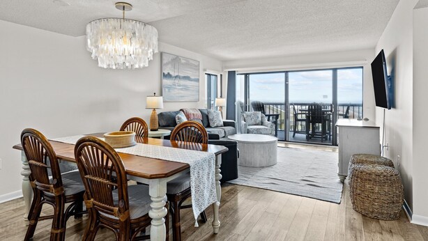 A spacious living room with a dining area, large windows, and a chandelier. - The Beach Hive: Ocean view condo in the heart of Emerald Isle (Emerald Isle)