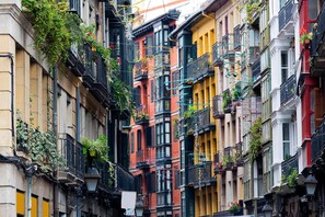 A narrow street with multi-story buildings, balconies with plants, and a mix of architectural styles.