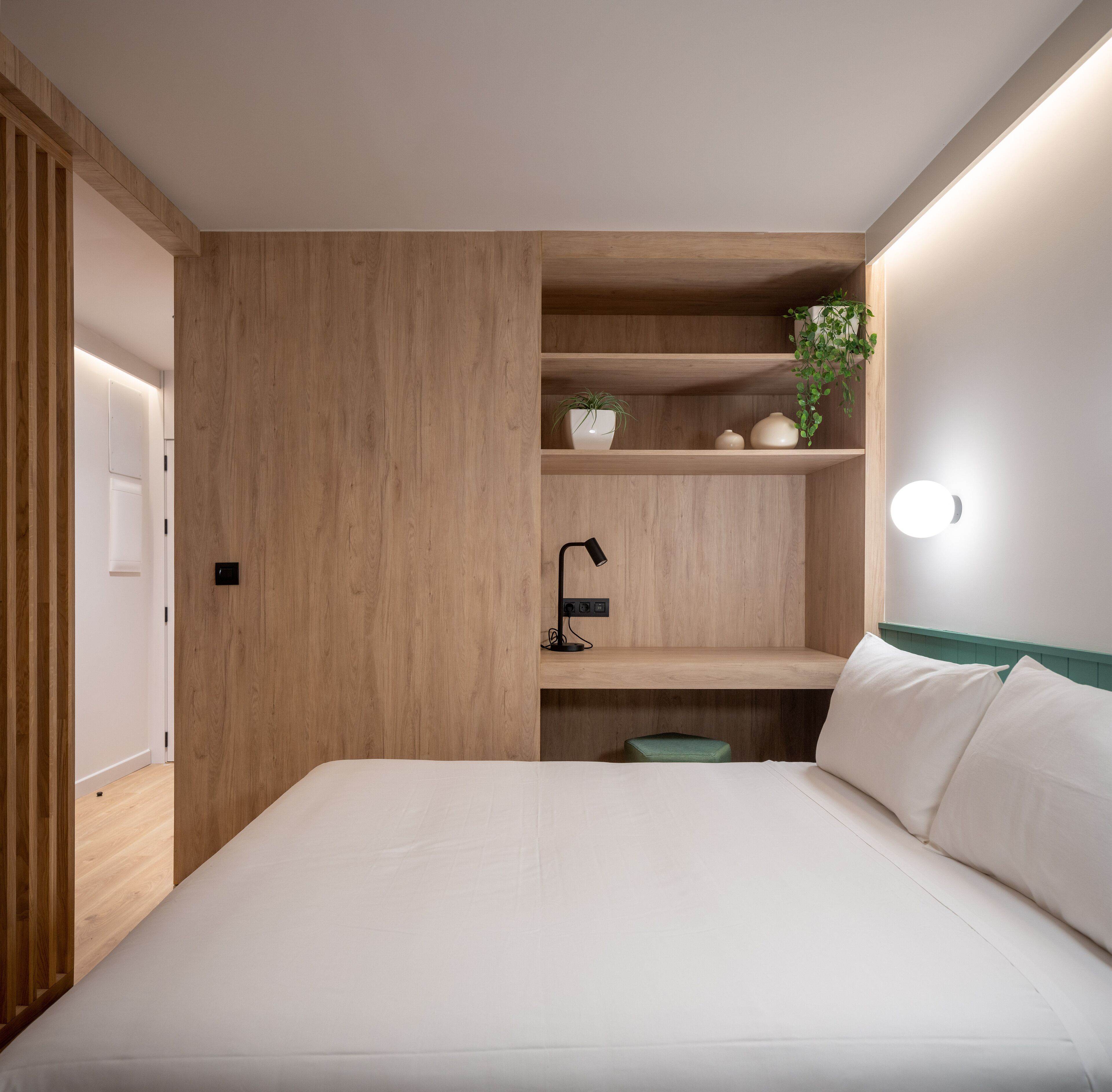 A modern bedroom with a wooden headboard, a bedside table with a lamp, and a shelf with decorative items.