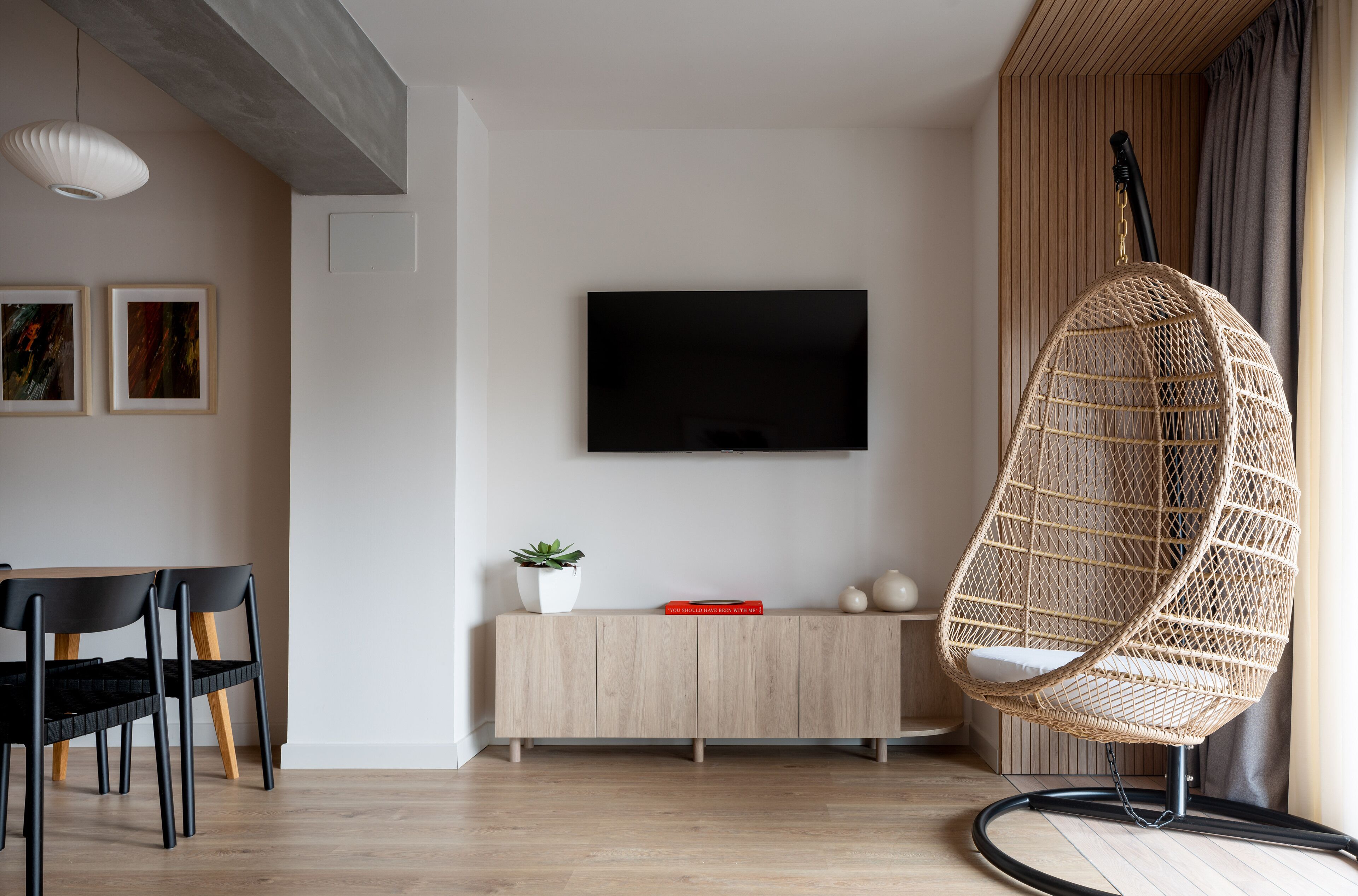Un soggiorno moderno con una TV a schermo piatto, un'area pranzo con un tavolo e sedie nere, una poltrona in vimini e un mobiletto in legno con una pianta.