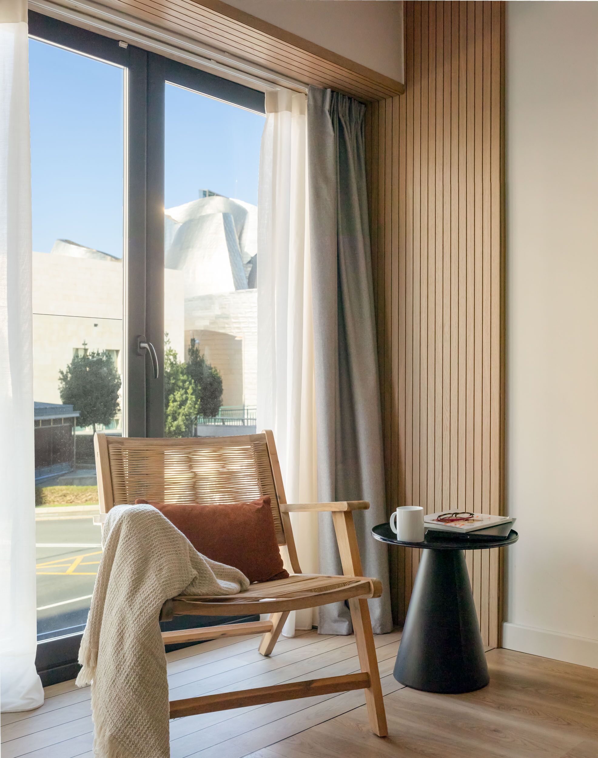 A modern room with a wooden chair, a small table with a cup and book, and a large window with curtains.