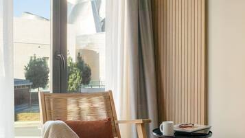 A modern room with a wooden chair, a small table with a cup and book, and a large window with curtains.