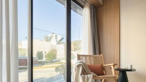 Une chambre moderne avec une grande fenêtre, un plancher en bois, une chaise en bois avec un coussin et une petite table ronde sur laquelle se trouve une tasse.