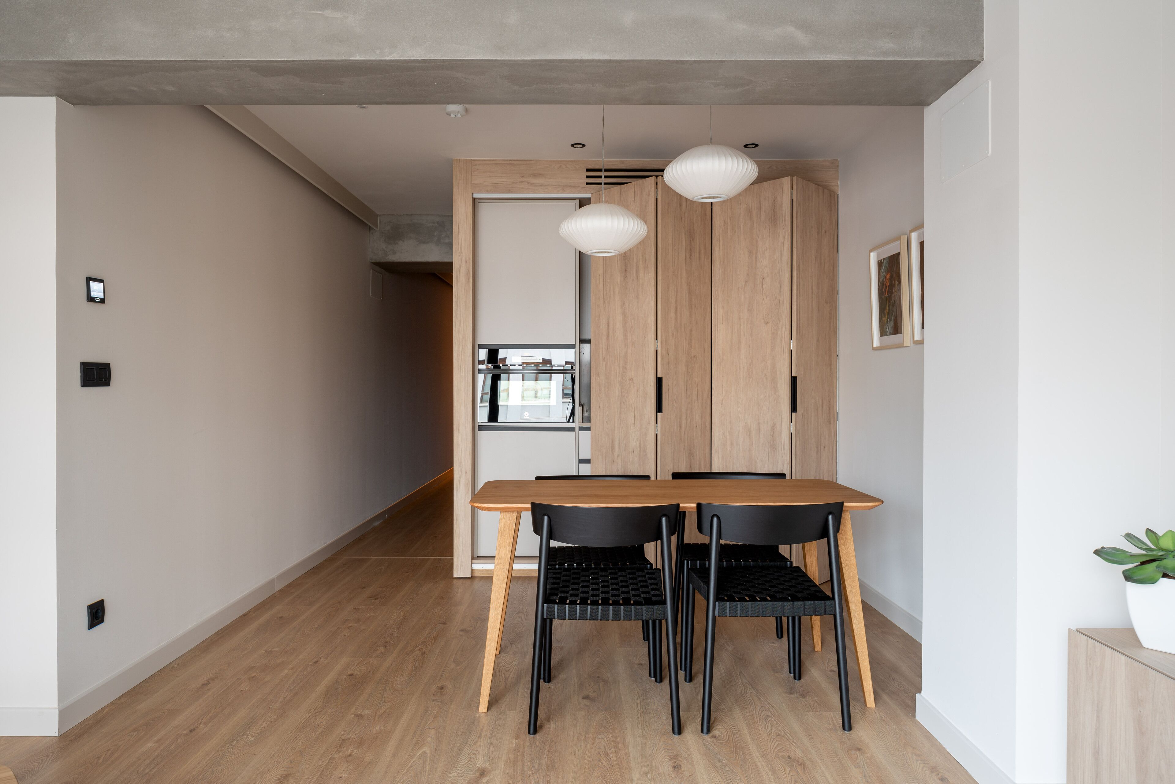 A modern dining area with a wooden table and chairs, a light fixture, and a view into the kitchen.