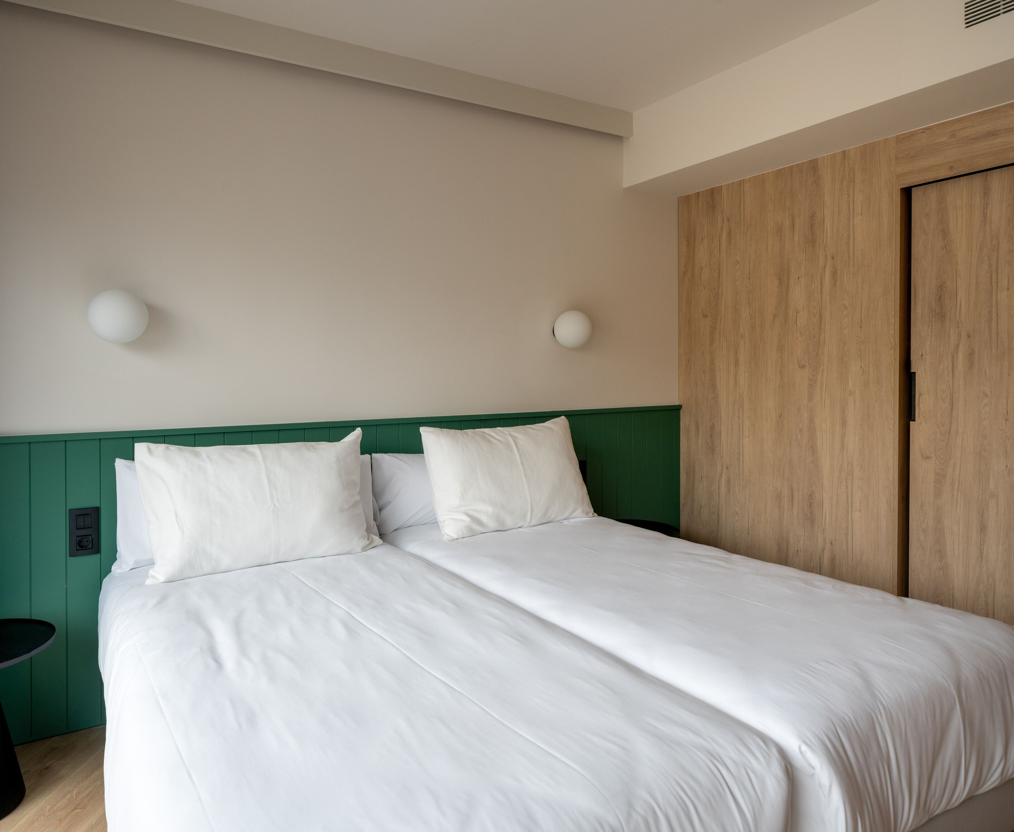 Un lit bien fait, avec du linge de lit blanc, dans une pièce aux murs recouverts de lambris verts et dotée d’une porte en bois.