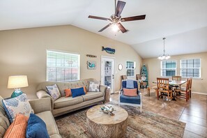 A spacious living room with a ceiling fan, a dining area with wooden chairs, and a kitchen visible through an open door. - Pirates Paradise:  Perfect Oasis! (Port Aransas)