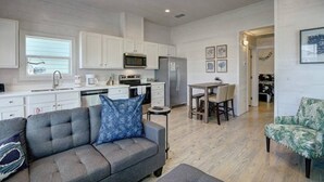 A modern kitchen with white cabinets, a dining area with a wooden table and chairs, and a gray sofa with a blue pillow.