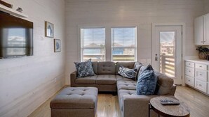 A cozy living room with a sectional sofa, ottoman, and a small round table. There are two windows with blinds, a door, and a kitchen area visible.