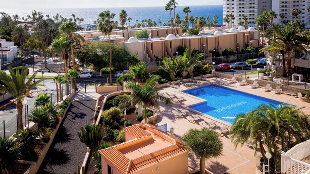 Un complex turístic amb piscina, palmeres i vista al mar.
