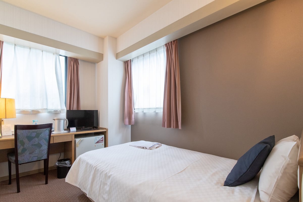 A hotel room with a bed, a desk with a television, a chair, and a window with curtains.
