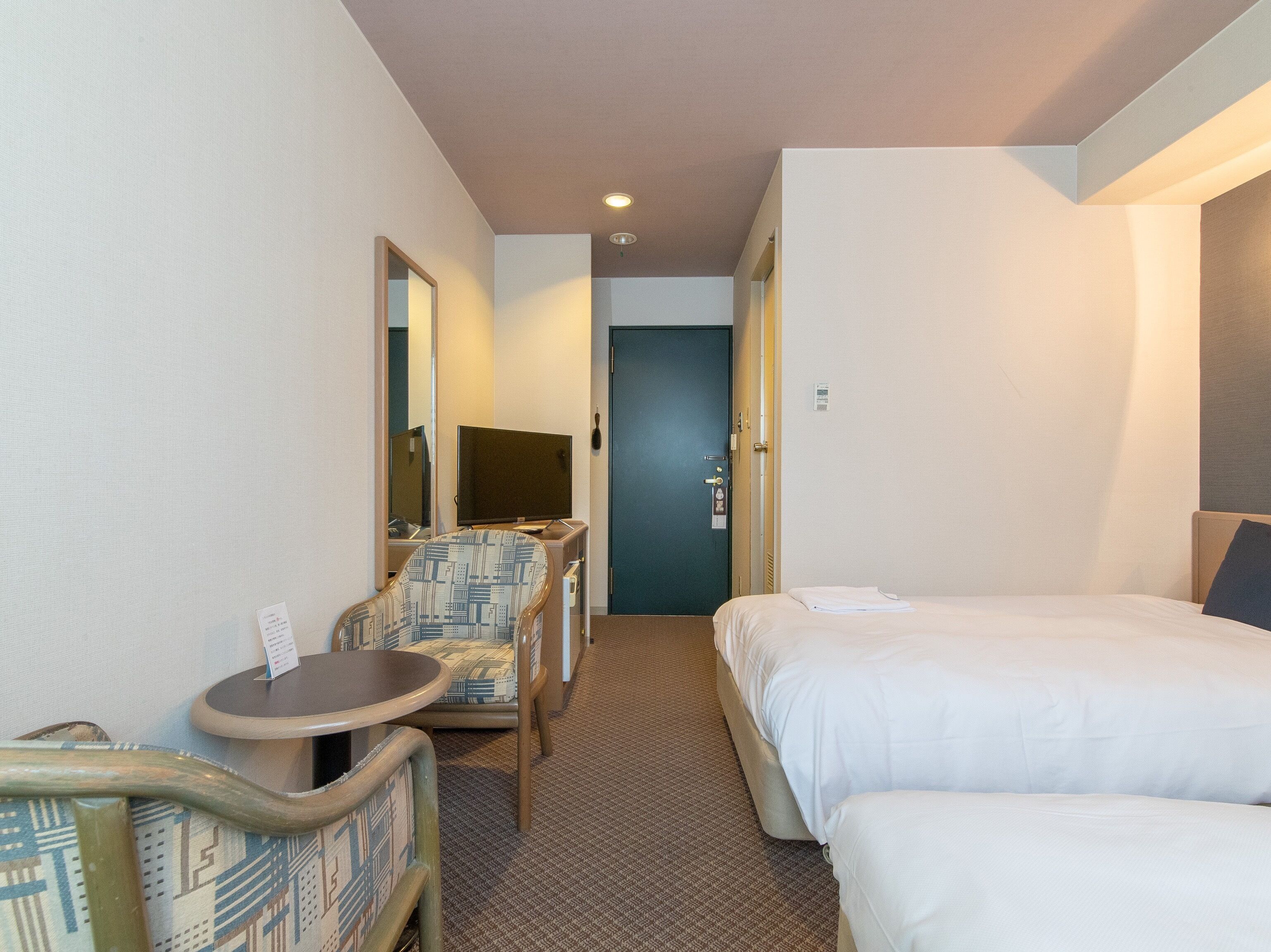 A hotel room with a bed, a chair, a small table, a mirror, and a door.