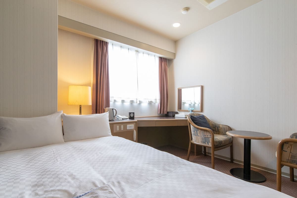 A hotel room with a large bed, a desk, a chair, a small table, a lamp, a mirror, and a window with curtains.