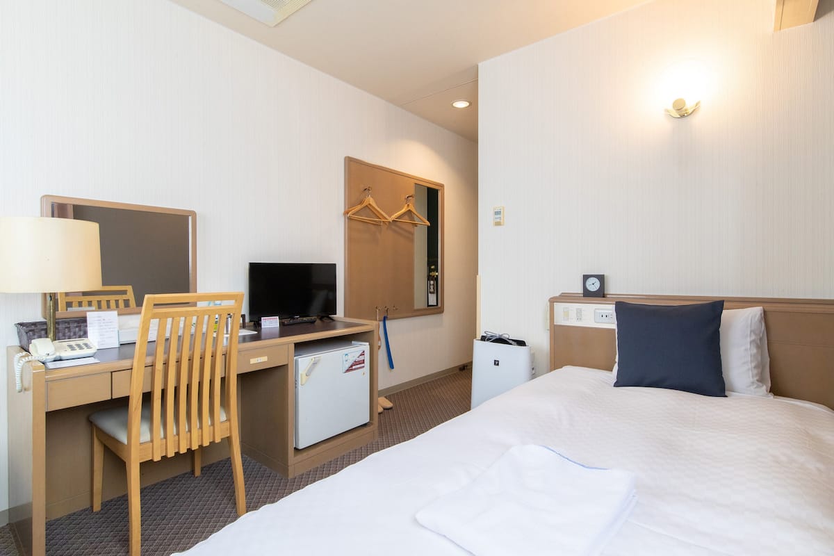 A hotel room with a bed, a desk with a computer, a chair, a TV, a small fridge, a bedside table, a lamp, and a clock on the wall.