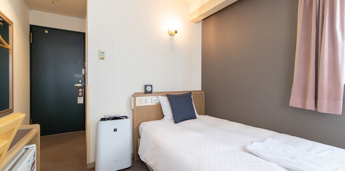 Quarto de hotel com uma cama, mesa de cabeceira, escrivaninha, cadeira, relógio na parede e uma porta.