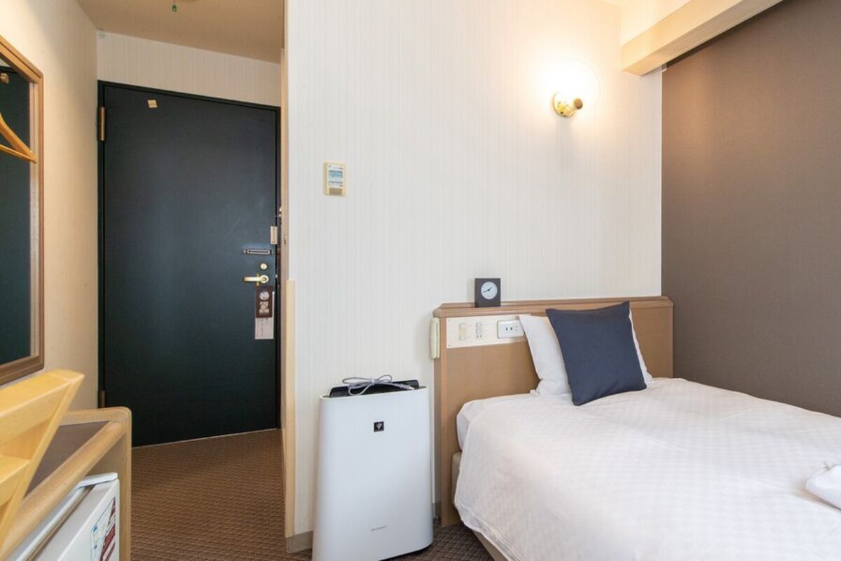 A hotel room with a bed, a bedside table, a small desk, a chair, a wall-mounted clock, and a door.