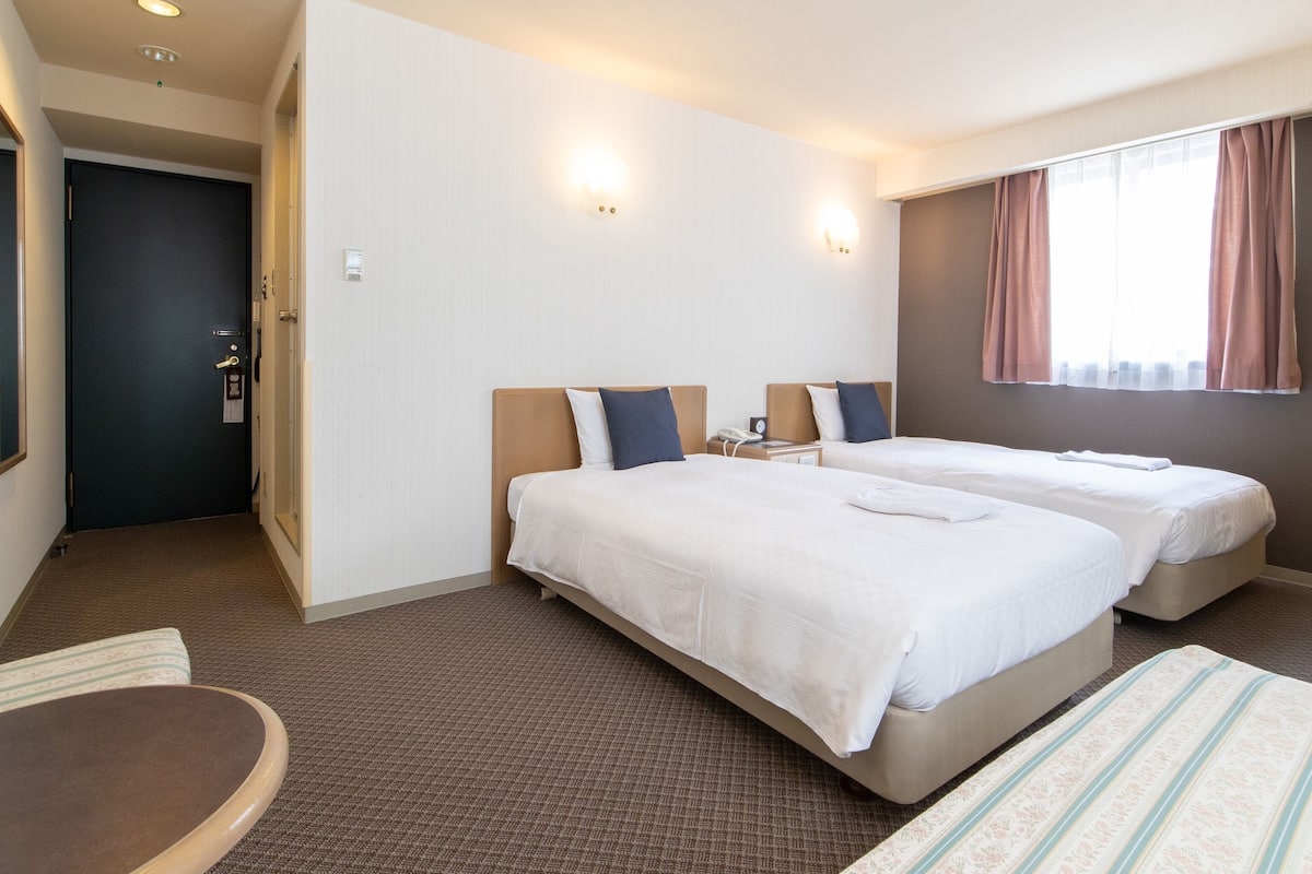 A hotel room with two beds, a desk, a chair, and a window with curtains.