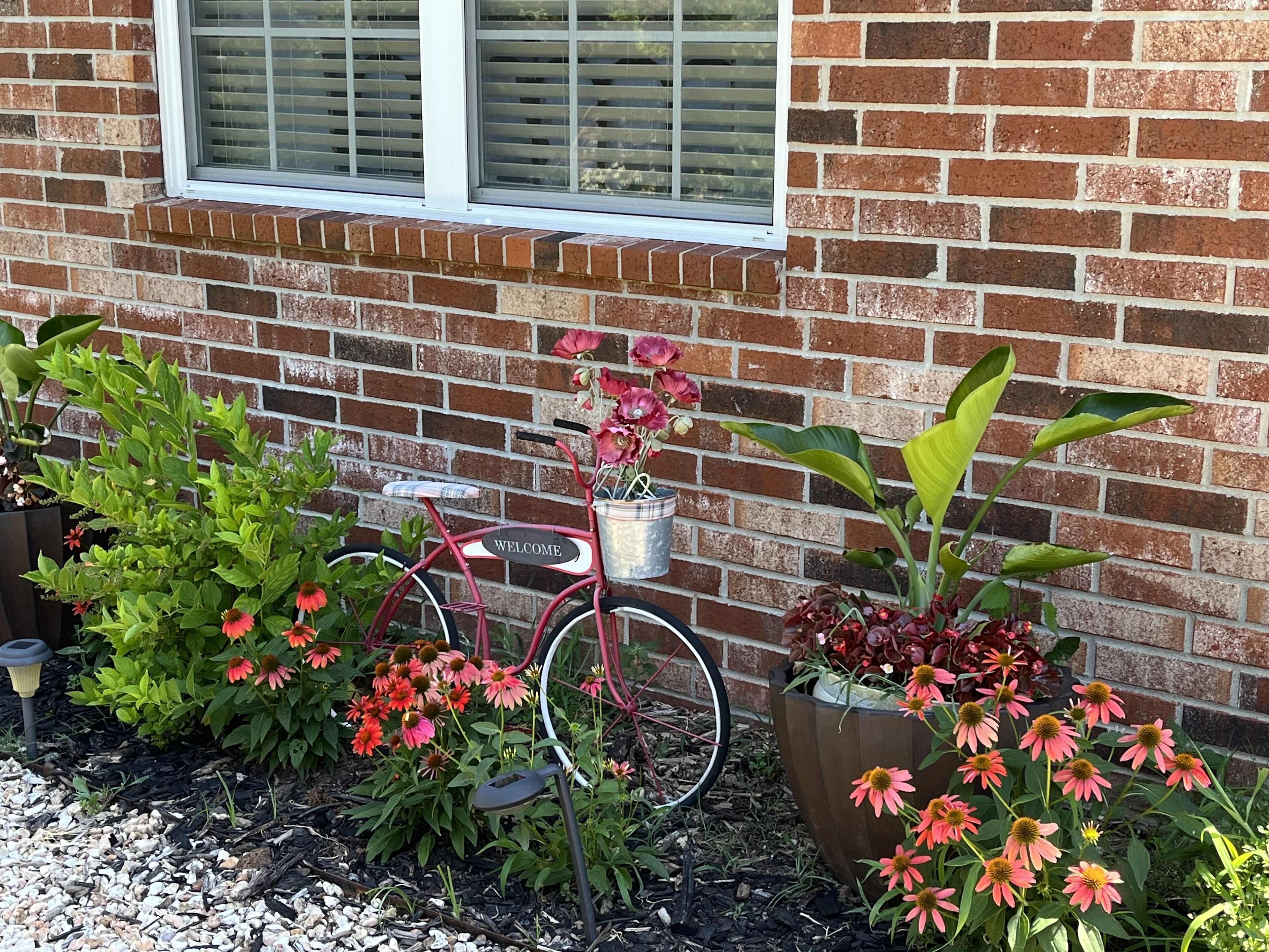 Ein rotes Fahrrad mit einem „WILLKOMMEN“-Schild, Blumentöpfen und einer Ziegelwand.