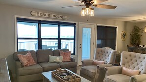 Een woonkamer met een plafondventilator, een bordje met daarop 'Beach House' en een uitzicht door grote ramen.