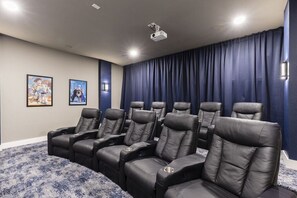 Una sala de cine en casa con seis butacas reclinables de cuero negro, una alfombra azul y un proyector en el techo.