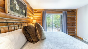 A bedroom with a wooden log wall, a bed with white and brown pillows, a nightstand with a lamp, and a view of the outdoors through a sliding glass door. - 57 Rasor Court by Summit County Mountain Retreats (Keystone)
