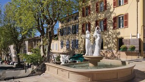 Une cour avec une fontaine, un arbre et un bâtiment aux volets rouges.