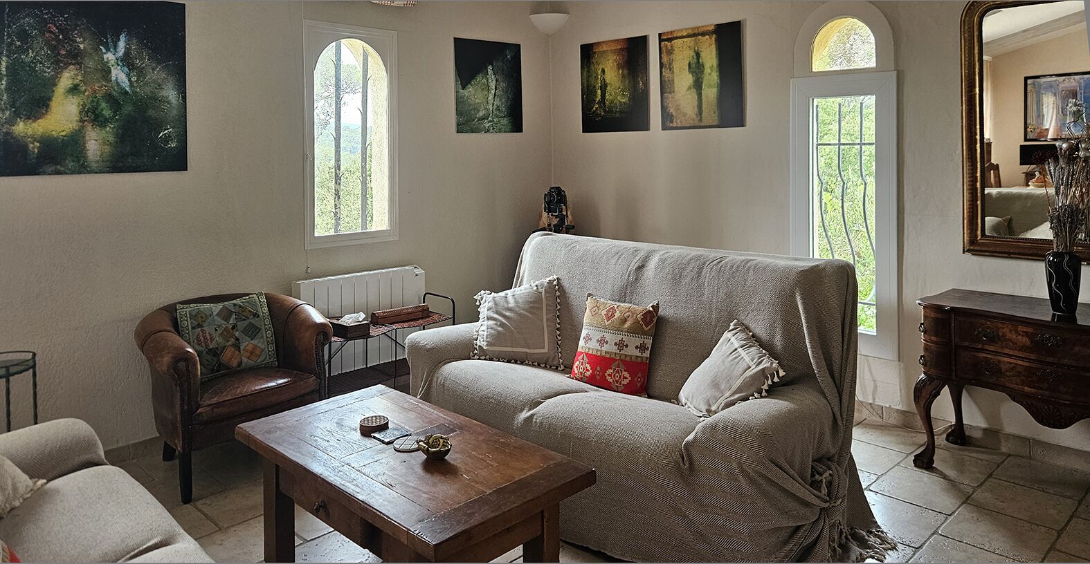 Ein gemütliches Wohnzimmer mit einer Couch, einem Sessel und einem Couchtisch. An den Wänden hängen Gemälde und auf der rechten Seite befindet sich ein Spiegel.