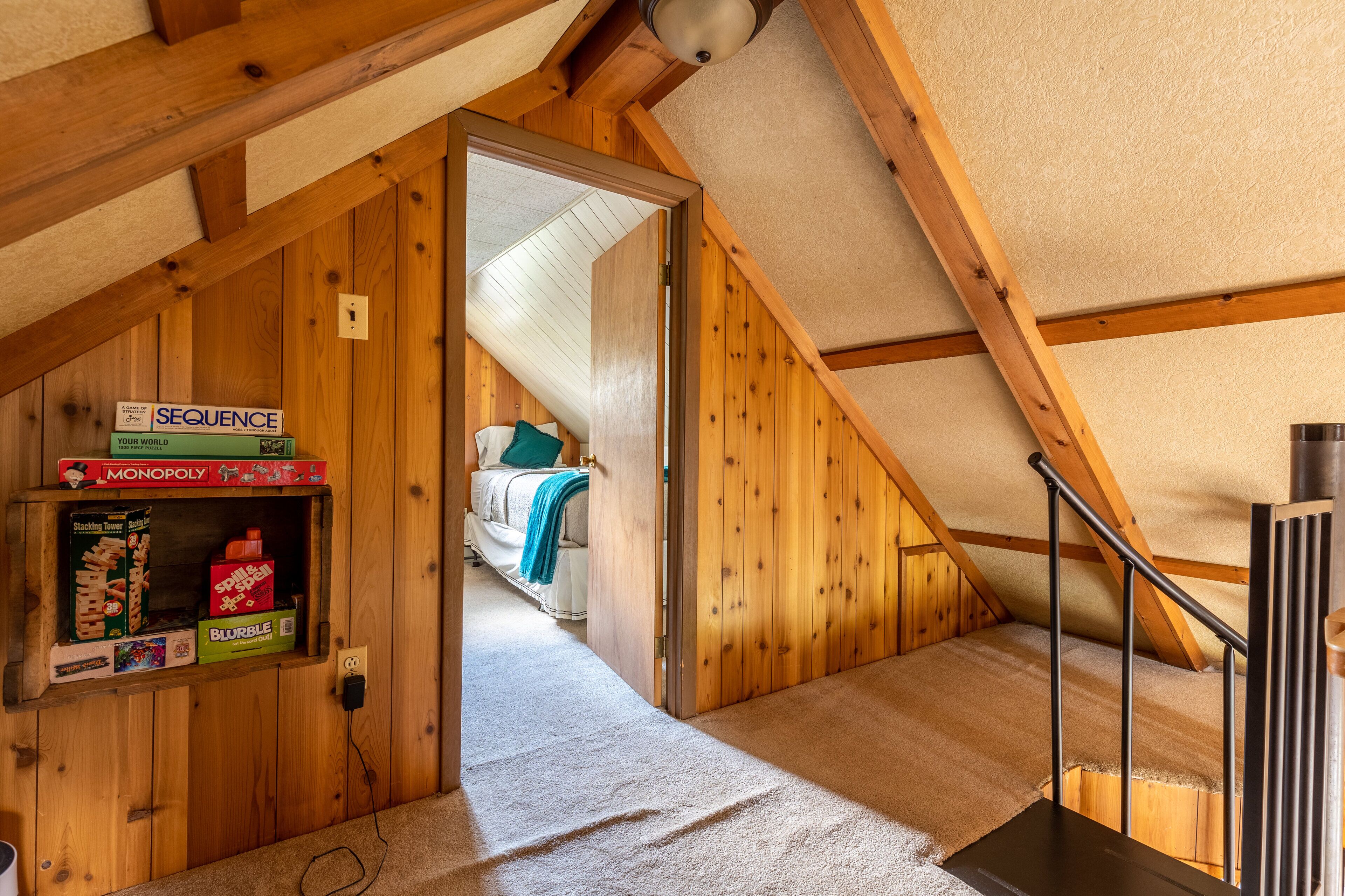 Een kamer met houten wanden, een schuine plafond, een bed met kussens, een plank met gezelschapsspellen en een vloerbedekking.
