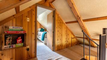 Een kamer met houten wanden, een schuine plafond, een bed met kussens, een plank met gezelschapsspellen en een vloerbedekking.