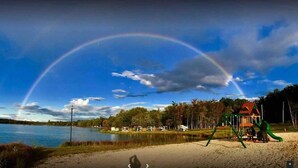 Vista de un lago con un arcoíris, un parque infantil y una cabaña al fondo.