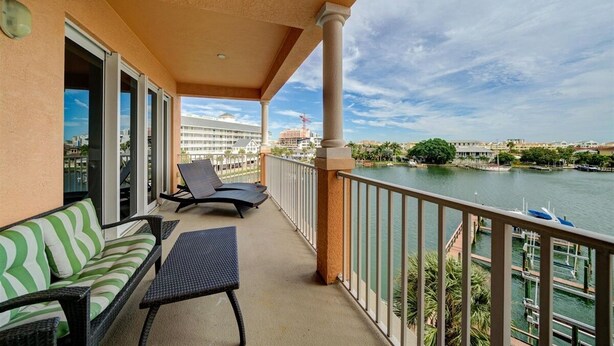 A balcony with a sofa, chairs, and a table overlooking a body of water with boats and buildings in the background.